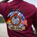 The image shows the back of a maroon/burgundy athletic shirt worn by a person, featuring a detailed military unit graphic. The shirt belongs to the 23rd MAOC (Munitions and Armaments Operations Company) and commemorates an 'Ammo Rodeo' event for FY26. The design incorporates the U.S. Army Ordnance Corps insignia flanked by German and American flags against a Bavarian blue-and-white diamond pattern background.