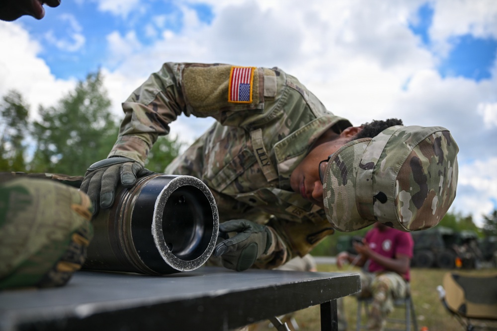A U.S. Army soldier in MultiCam/OCP camouflage uniform is bent over a field table closely inspecting or working on a cylindrical military equipment component, likely a mortar tube or launcher barrel. The soldier wears tactical gloves and a patrol cap with a U.S. flag patch visible on the right sleeve. The setting appears to be an outdoor field training or exercise environment.