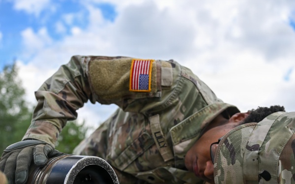 A U.S. Army soldier in MultiCam/OCP camouflage uniform is bent over a field table closely inspecting or working on a cylindrical military equipment component, likely a mortar tube or launcher barrel. The soldier wears tactical gloves and a patrol cap with a U.S. flag patch visible on the right sleeve. The setting appears to be an outdoor field training or exercise environment.