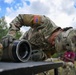 A U.S. Army soldier in MultiCam/OCP camouflage uniform is bent over a field table closely inspecting or working on a cylindrical military equipment component, likely a mortar tube or launcher barrel. The soldier wears tactical gloves and a patrol cap with a U.S. flag patch visible on the right sleeve. The setting appears to be an outdoor field training or exercise environment.
