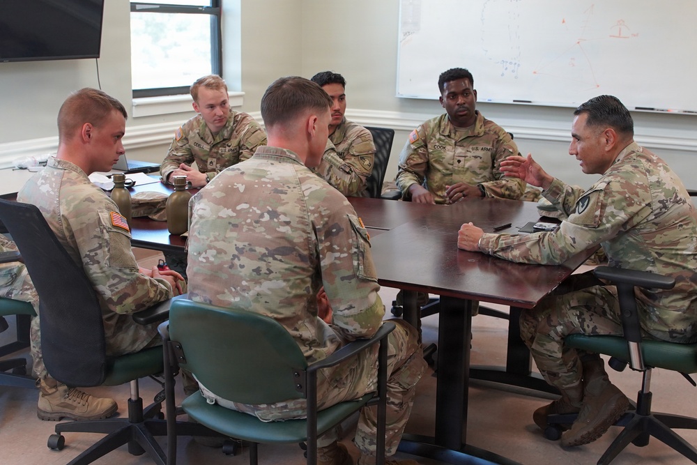 A group of six U.S. Army soldiers in Operational Camouflage Pattern (OCP) uniforms are seated around a conference table in what appears to be a military briefing or meeting room. One soldier on the right side of the table appears to be leading the discussion, with others listening attentively. A whiteboard with diagrams is visible in the background.