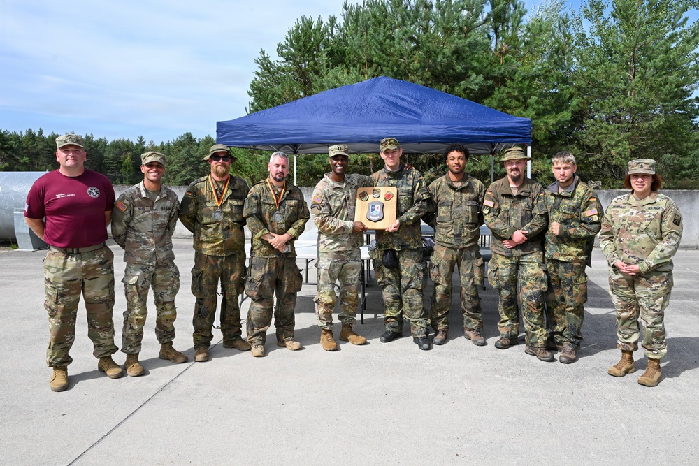 A group of approximately ten military personnel pose for a formal group photograph outdoors near a blue canopy tent. Two central figures are holding a wooden plaque or award shield displaying multiple unit patches or insignia. The group appears to include both U.S. Army soldiers in OCP (Operational Camouflage Pattern) uniforms and German Bundeswehr soldiers in Flecktarn camouflage, suggesting a joint military event or competition.