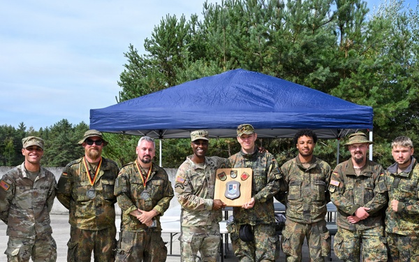 A group of approximately ten military personnel pose for a formal group photograph outdoors near a blue canopy tent. Two central figures are holding a wooden plaque or award shield displaying multiple unit patches or insignia. The group appears to include both U.S. Army soldiers in OCP (Operational Camouflage Pattern) uniforms and German Bundeswehr soldiers in Flecktarn camouflage, suggesting a joint military event or competition.