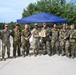 A group of approximately ten military personnel pose for a formal group photograph outdoors near a blue canopy tent. Two central figures are holding a wooden plaque or award shield displaying multiple unit patches or insignia. The group appears to include both U.S. Army soldiers in OCP (Operational Camouflage Pattern) uniforms and German Bundeswehr soldiers in Flecktarn camouflage, suggesting a joint military event or competition.