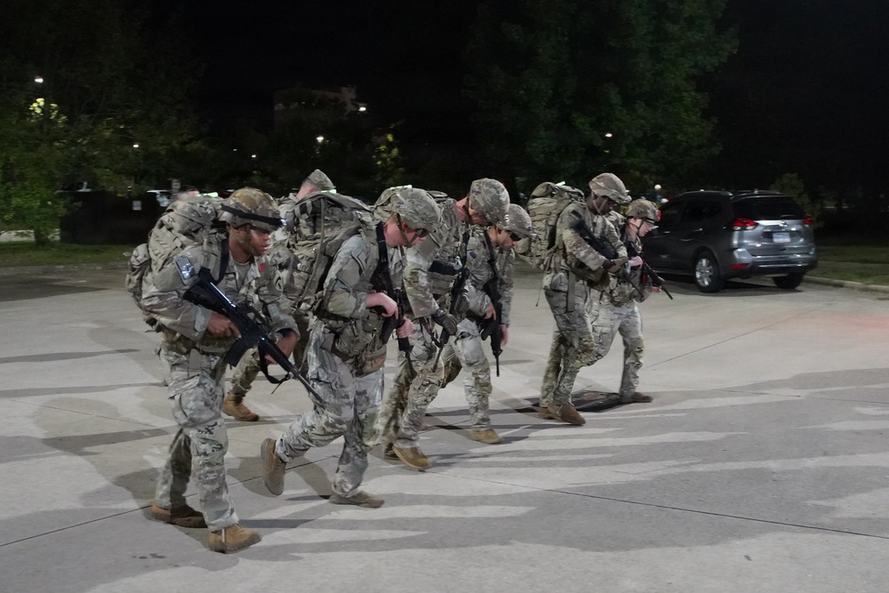 A group of military personnel in full combat gear are conducting a ruck march at night in what appears to be a parking lot or paved area. The soldiers are wearing camouflage uniforms, carrying large rucksacks, and armed with rifles. The setting appears to be a training exercise or operational movement based on the organized formation and equipment.