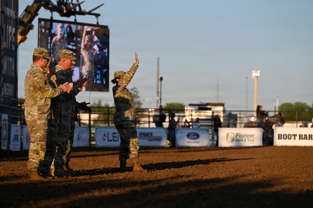 Three military personnel in camouflage uniforms are standing in a rodeo arena dirt floor during what appears to be a recognition or ceremony event. One service member is waving to the crowd while the others applaud. The setting is an outdoor rodeo venue with sponsor banners, a large video screen, and arena fencing visible in the background.