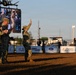 Three military personnel in camouflage uniforms are standing in a rodeo arena dirt floor during what appears to be a recognition or ceremony event. One service member is waving to the crowd while the others applaud. The setting is an outdoor rodeo venue with sponsor banners, a large video screen, and arena fencing visible in the background.