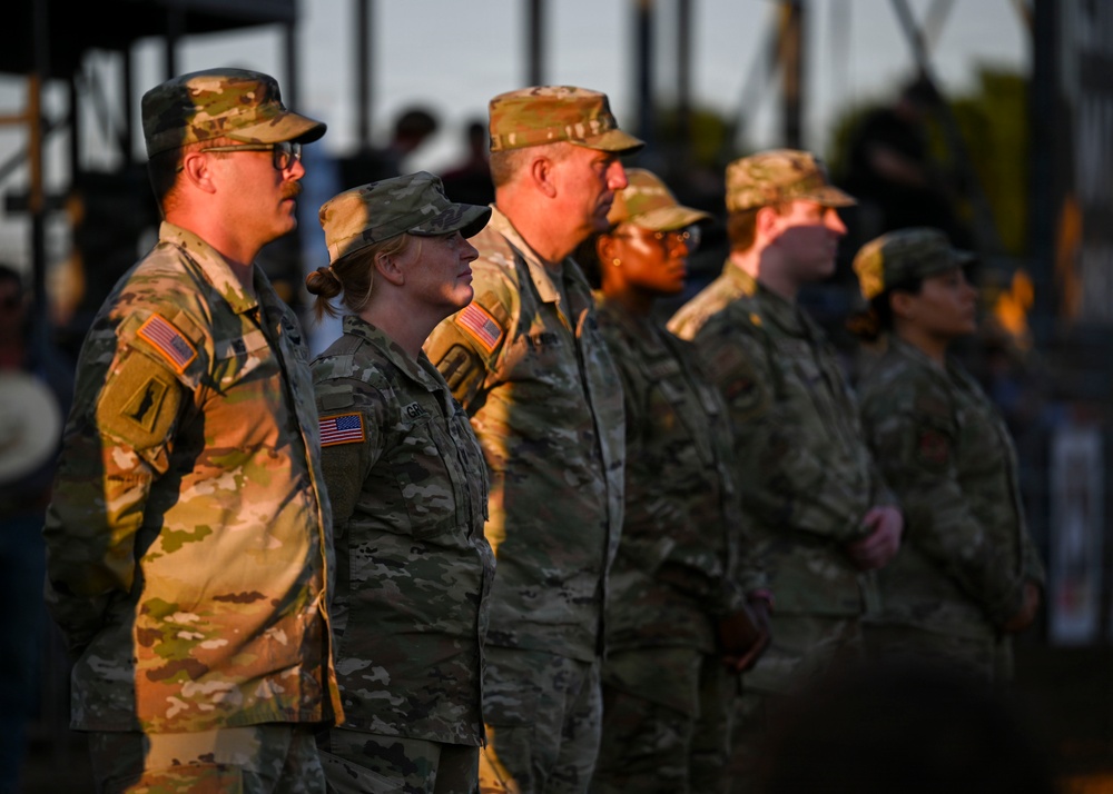 A group of U.S. Army soldiers in Operational Camouflage Pattern (OCP) uniforms are standing in formation at what appears to be an outdoor ceremony or event. The soldiers include both male and female personnel, with American flag patches visible on their left shoulders. The scene is lit by warm golden-hour sunlight, suggesting either early morning or late afternoon.