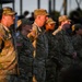 A group of U.S. Army soldiers in Operational Camouflage Pattern (OCP) uniforms are standing in formation at what appears to be an outdoor ceremony or event. The soldiers include both male and female personnel, with American flag patches visible on their left shoulders. The scene is lit by warm golden-hour sunlight, suggesting either early morning or late afternoon.