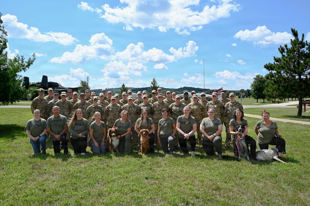 A large group photo of military personnel in camouflage uniforms standing in the back rows, with civilian handlers wearing matching olive green 'OPK9' t-shirts kneeling in the front row alongside several dogs. The setting appears to be a military installation or airfield, with what appears to be a vintage aircraft visible in the background. This appears to be an Operation K9 (OPK9) event or training program connecting military members with service or therapy dogs.