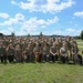 A large group photo of military personnel in camouflage uniforms standing in the back rows, with civilian handlers wearing matching olive green 'OPK9' t-shirts kneeling in the front row alongside several dogs. The setting appears to be a military installation or airfield, with what appears to be a vintage aircraft visible in the background. This appears to be an Operation K9 (OPK9) event or training program connecting military members with service or therapy dogs.
