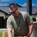 A military personnel member wearing an olive drab t-shirt and multicam/OCP pattern pants is attending to a military working dog (black in color) outdoors. He is applying what appears to be a bandage or gauze to the dog while a tactical medical kit is open nearby. The setting appears to be a military installation during daylight hours.