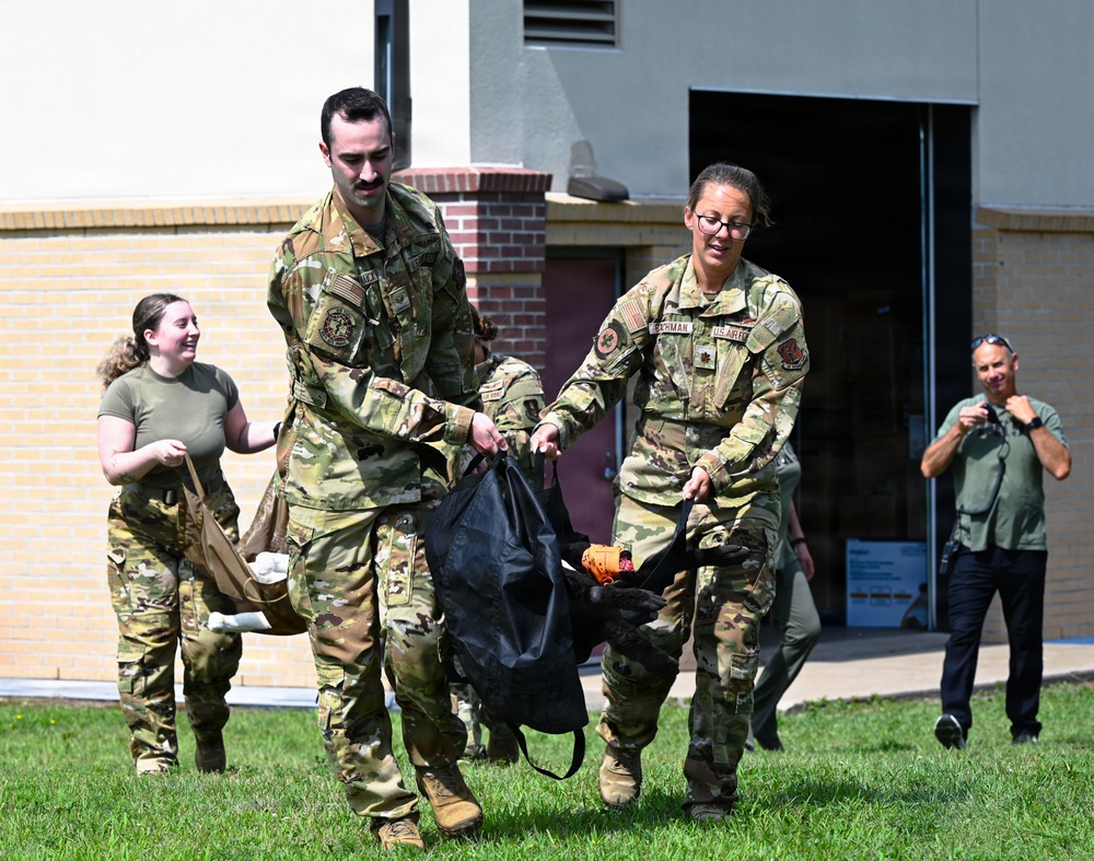 Two U.S. Air Force personnel in OCP (Operational Camouflage Pattern) uniforms are carrying a black bag together across a grassy area outside a brick building. A third service member in the background carries additional gear, while a civilian in a green shirt observes and appears to be photographing the activity. The scene appears to be a training exercise or equipment movement drill at a military installation.