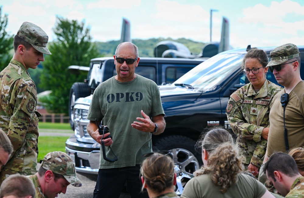 A civilian instructor wearing an 'OPK9' t-shirt addresses a group of military personnel in MultiCam/OCP uniforms outdoors. The group appears to be receiving a briefing or instruction, with some personnel seated and others standing attentively. A female U.S. Army officer with a rank insignia visible on her uniform is among those listening.