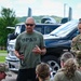 A civilian instructor wearing an 'OPK9' t-shirt addresses a group of military personnel in MultiCam/OCP uniforms outdoors. The group appears to be receiving a briefing or instruction, with some personnel seated and others standing attentively. A female U.S. Army officer with a rank insignia visible on her uniform is among those listening.