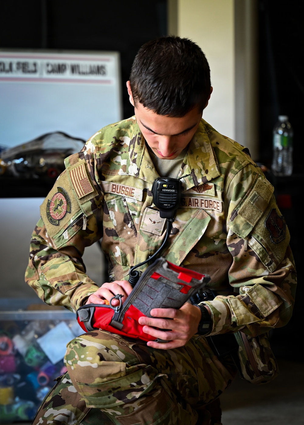 A U.S. Air Force service member named Bussie, wearing OCP (Operational Camouflage Pattern) uniform, is seated and examining what appears to be a red medical or tactical pouch. He has a Kenwood KMC-41 speaker-microphone clipped to his chest, indicating active radio communication capability. The background shows a sign partially reading 'Camp Williams' and various equipment.