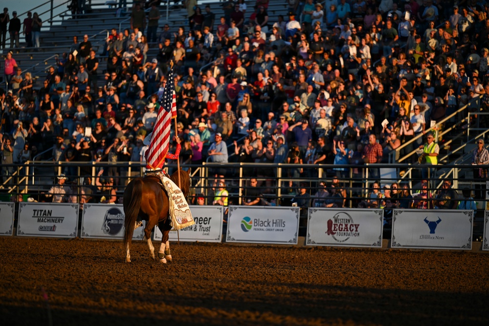 A rodeo arena scene featuring a rider on a chestnut horse carrying an American flag, likely during an opening ceremony or grand entry. The packed grandstands are filled with hundreds of spectators, many appearing to applaud or stand in respect. Sponsor banners line the arena fence, and the scene is bathed in warm golden evening light.