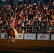 A rodeo arena scene featuring a rider on a chestnut horse carrying an American flag, likely during an opening ceremony or grand entry. The packed grandstands are filled with hundreds of spectators, many appearing to applaud or stand in respect. Sponsor banners line the arena fence, and the scene is bathed in warm golden evening light.