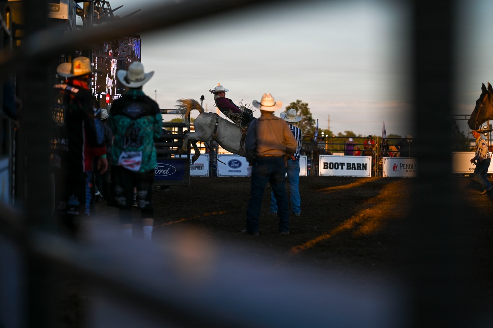 A rodeo event photographed through metal fence rails at dusk or early evening, showing a bucking horse with a rider in the arena. Sponsors including Boot Barn, Ford, and Pendleton are visible on arena banners. Several cowboys and rodeo personnel are present both inside and outside the arena.