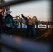 A rodeo event photographed through metal fence rails at dusk or early evening, showing a bucking horse with a rider in the arena. Sponsors including Boot Barn, Ford, and Pendleton are visible on arena banners. Several cowboys and rodeo personnel are present both inside and outside the arena.
