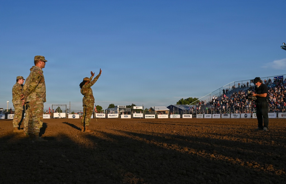 Three U.S. Army soldiers in OCP (Operational Camouflage Pattern) uniforms are standing in a rodeo arena, with one soldier raising both hands, possibly addressing the crowd or being recognized. A civilian with a camera is filming or photographing them from the right side. The grandstands in the background are filled with spectators, and sponsor banners including 'CINCH,' 'BLAKE'S,' and 'RESISTOL' are visible along the arena fence.