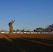 Three U.S. Army soldiers in OCP (Operational Camouflage Pattern) uniforms are standing in a rodeo arena, with one soldier raising both hands, possibly addressing the crowd or being recognized. A civilian with a camera is filming or photographing them from the right side. The grandstands in the background are filled with spectators, and sponsor banners including 'CINCH,' 'BLAKE'S,' and 'RESISTOL' are visible along the arena fence.