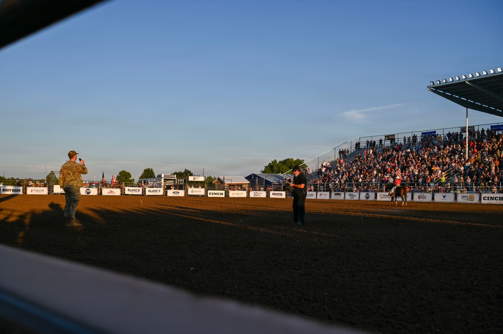 A rodeo arena event captured at golden hour, showing a packed grandstand of spectators watching an opening ceremony or performance. A person in military camouflage uniform stands on the left side of the arena holding a microphone, while another person in black stands near the center. Sponsor banners including Boot Barn, Cinch, Ford, and Blake's line the arena fence.