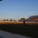 A rodeo arena event captured at golden hour, showing a packed grandstand of spectators watching an opening ceremony or performance. A person in military camouflage uniform stands on the left side of the arena holding a microphone, while another person in black stands near the center. Sponsor banners including Boot Barn, Cinch, Ford, and Blake's line the arena fence.