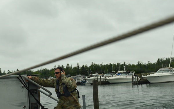 A military personnel in camouflage uniform is standing on the stern of a military vessel at a marina, appearing to assist with docking or departure operations. The individual is wearing a life vest, sunglasses, and hearing protection while gripping a railing near a dock. Several civilian recreational boats are visible in the background marina.