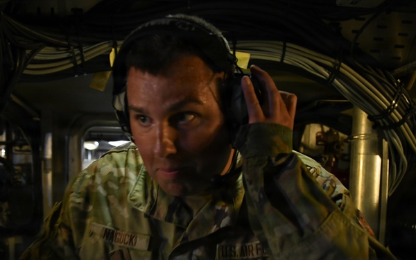 A U.S. Air Force service member in OCP (Operational Camouflage Pattern) uniform is photographed inside what appears to be a military aircraft, likely a large transport or tanker. The individual is wearing headphones/headset and appears to be listening or communicating while navigating through a narrow interior passage. The environment is dimly lit with visible cables, tubing, and mechanical components characteristic of a military aircraft interior.