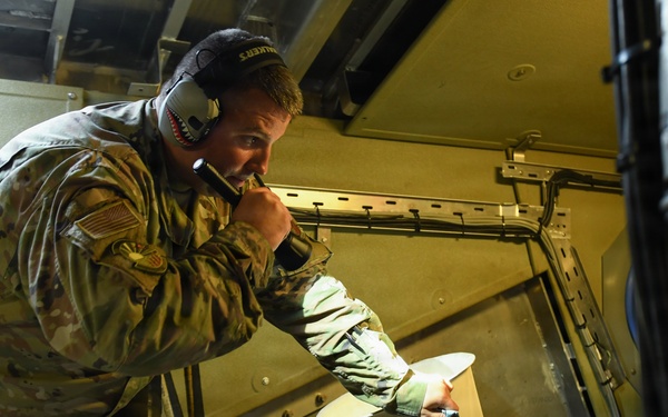 A U.S. Army soldier in MultiCam/OCP camouflage uniform is working inside what appears to be a military vehicle or aircraft interior. The soldier is wearing communications headgear and appears to be speaking into a microphone while handling or inspecting equipment. The setting suggests a technical or operational maintenance/communications task.
