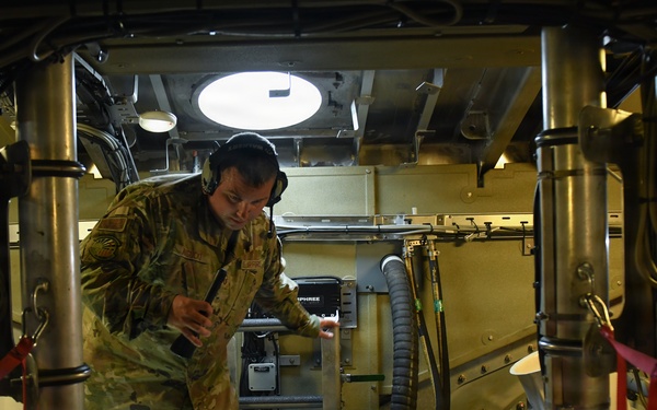 A soldier in camouflage uniform and communications headset is working inside what appears to be a military vehicle or aircraft cargo bay, operating or inspecting equipment. The interior is densely packed with electronic systems, wiring, control panels, and mechanical components secured with red cargo straps. The environment suggests a technical maintenance or operational check is being performed on specialized military equipment.
