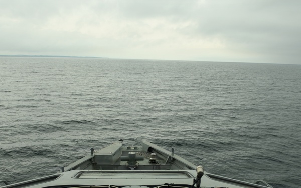 The image shows the forward bow deck of a military or naval vessel underway on calm, overcast open water. The deck features various mounted equipment including what appears to be a gun mount base ring, cable bundles, and electronic equipment housings. A distant shoreline is faintly visible on the horizon under grey, cloudy skies.