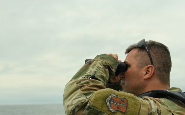 A military personnel member in MultiCam/OCP camouflage uniform is standing on what appears to be the deck of a naval vessel, scanning the open sea with binoculars. The individual is wearing a life vest over their uniform and has sunglasses resting on their head. The overcast sky and calm open water form the backdrop of this maritime surveillance scene.