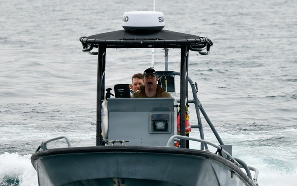 A gray military or law enforcement patrol boat is moving through open ocean waters toward the camera. Two personnel are visible at the helm station, which is equipped with a radar dome and navigation electronics. The vessel is creating a visible bow wake, indicating it is underway at moderate speed.