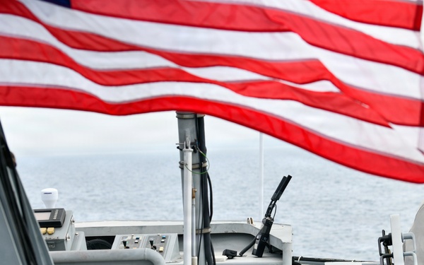 The image shows a U.S. military or Coast Guard vessel underway on open water, with an American flag prominently flying in the foreground. At least two personnel in military/tactical uniforms are visible inside what appears to be the bridge or command station of the vessel. Various navigation and communication equipment are visible on the deck.