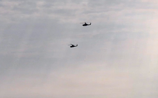 The image shows a maritime scene with two helicopters flying in formation over open water. Below them, at least two watercraft are visible on the surface, one of which appears to be moving at speed and creating a wake. The overcast sky and hazy conditions suggest either early morning or overcast weather, and the overall scene has the appearance of a military or coast guard operation.