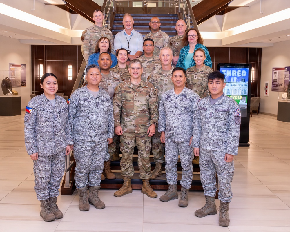 A formal group photo of approximately 16 military personnel and civilians posed on and in front of a staircase in what appears to be a modern military facility lobby. The group includes members wearing U.S. Air Force OCP (Operational Camouflage Pattern) uniforms and Philippine Air Force digital camouflage uniforms, along with several civilians in business casual attire. Name tapes visible on some Philippine Air Force members include 'MANALO,' 'MARA,' 'ATD,' and 'ROSELE,' suggesting a bilateral or exchange military engagement.