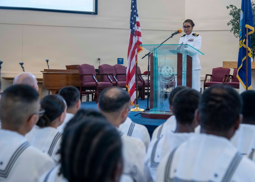 A U.S. Navy officer in dress white uniform is delivering a speech at a podium during a formal military ceremony. The audience consists of Navy enlisted personnel seated in rows, also wearing white uniforms. The setting appears to be a military chapel or ceremonial hall, with an American flag and a Navy flag flanking the speaker.