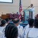 A U.S. Navy officer in dress white uniform is delivering a speech at a podium during a formal military ceremony. The audience consists of Navy enlisted personnel seated in rows, also wearing white uniforms. The setting appears to be a military chapel or ceremonial hall, with an American flag and a Navy flag flanking the speaker.
