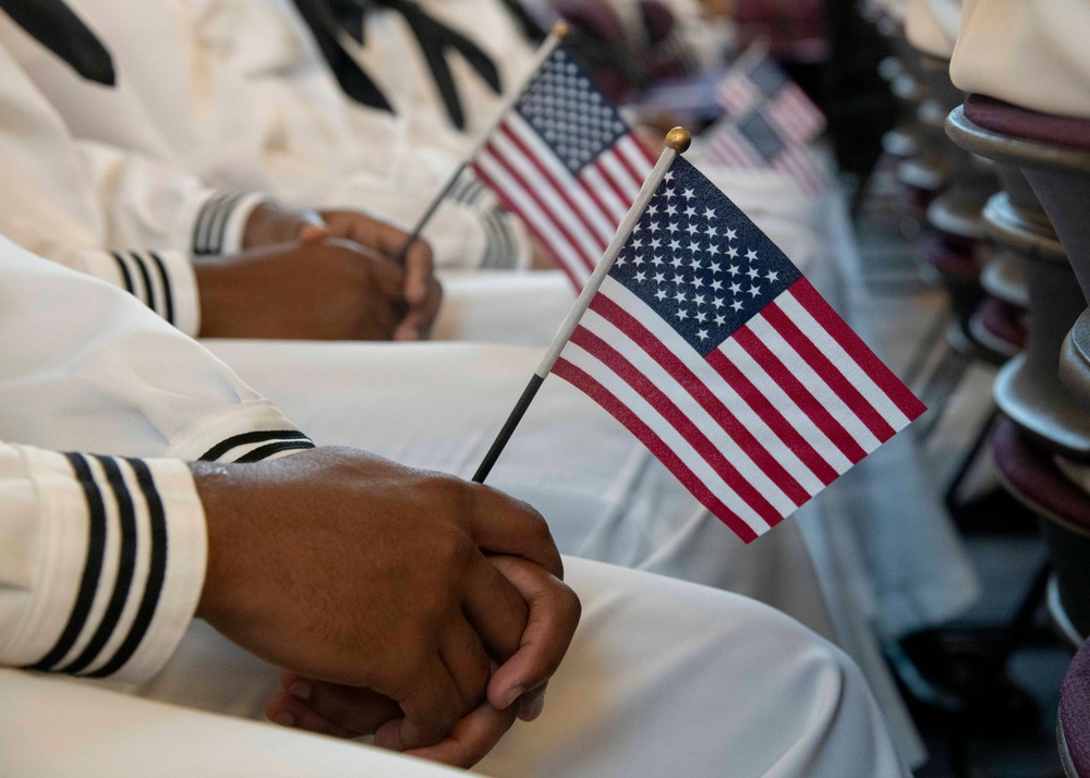 The image shows individuals in white military-style uniforms seated and holding small American flags. The focus is on their hands and laps, with the flags prominently displayed in the foreground. The setting appears to be a formal military ceremony or official event.
