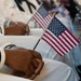 The image shows individuals in white military-style uniforms seated and holding small American flags. The focus is on their hands and laps, with the flags prominently displayed in the foreground. The setting appears to be a formal military ceremony or official event.