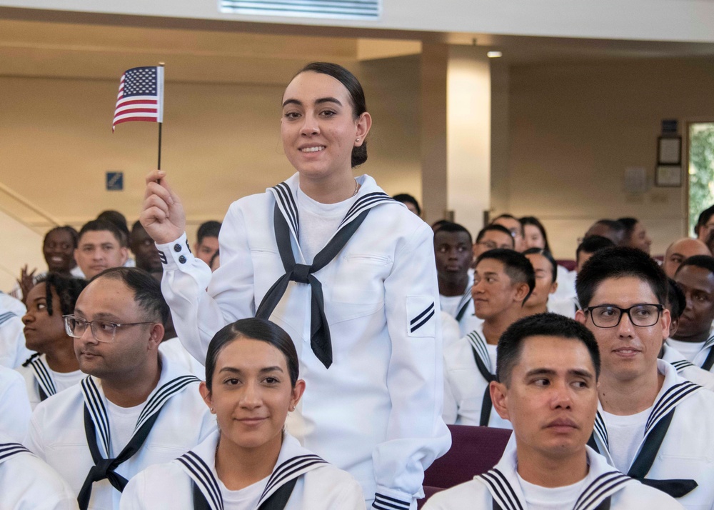 A group of U.S. Navy sailors in white dress uniforms are seated in what appears to be a ceremony hall. One female sailor is standing and waving a small American flag, smiling at the camera, suggesting this is a celebratory or official ceremony such as a naturalization or graduation event. The crowd behind her shows a diverse group of sailors also in uniform.