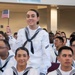 A group of U.S. Navy sailors in white dress uniforms are seated in what appears to be a ceremony hall. One female sailor is standing and waving a small American flag, smiling at the camera, suggesting this is a celebratory or official ceremony such as a naturalization or graduation event. The crowd behind her shows a diverse group of sailors also in uniform.