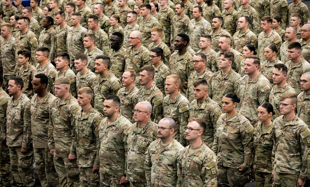 A large formation of U.S. Army soldiers standing at attention in what appears to be an indoor military ceremony or formation event. All personnel are wearing the Army Combat Uniform (ACU) in Operational Camouflage Pattern (OCP). The formation consists of both male and female soldiers arranged in multiple rows extending deep into the background.