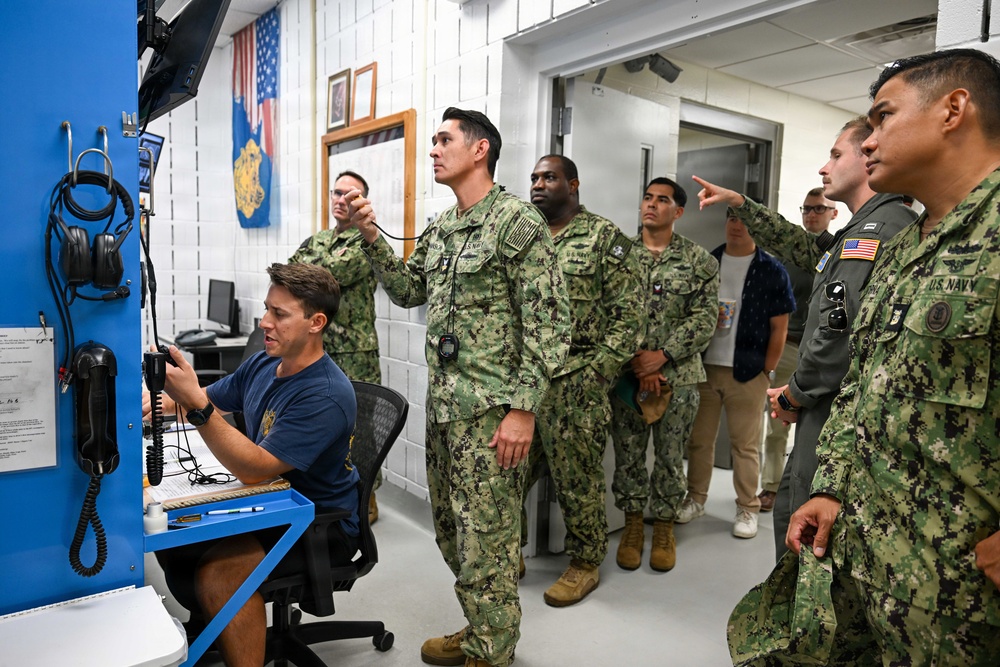 A group of U.S. Navy personnel in NWU Type III camouflage uniforms are gathered in what appears to be a military operations or communications room. A seated individual in a blue Navy t-shirt is operating communications equipment at a workstation while a standing officer points toward a monitor or display. The scene suggests a briefing, demonstration, or operational walkthrough at a naval facility.