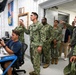 A group of U.S. Navy personnel in NWU Type III camouflage uniforms are gathered in what appears to be a military operations or communications room. A seated individual in a blue Navy t-shirt is operating communications equipment at a workstation while a standing officer points toward a monitor or display. The scene suggests a briefing, demonstration, or operational walkthrough at a naval facility.