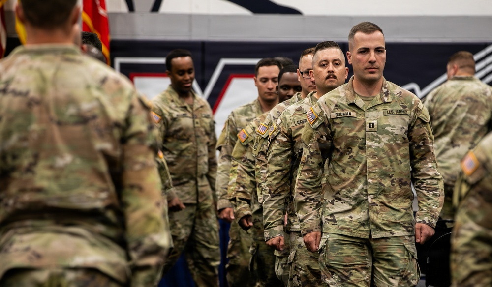 A group of U.S. Army soldiers in OCP (Operational Camouflage Pattern) uniforms are participating in a formal military ceremony, likely a deployment or unit activation event held indoors. The lead soldier, identified by name tape as 'BOUMAN,' wears a Captain's rank insignia and is marching in formation. The setting appears to be a gymnasium or arena with patriotic wall graphics visible in the background.