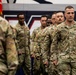 A group of U.S. Army soldiers in OCP (Operational Camouflage Pattern) uniforms are participating in a formal military ceremony, likely a deployment or unit activation event held indoors. The lead soldier, identified by name tape as 'BOUMAN,' wears a Captain's rank insignia and is marching in formation. The setting appears to be a gymnasium or arena with patriotic wall graphics visible in the background.