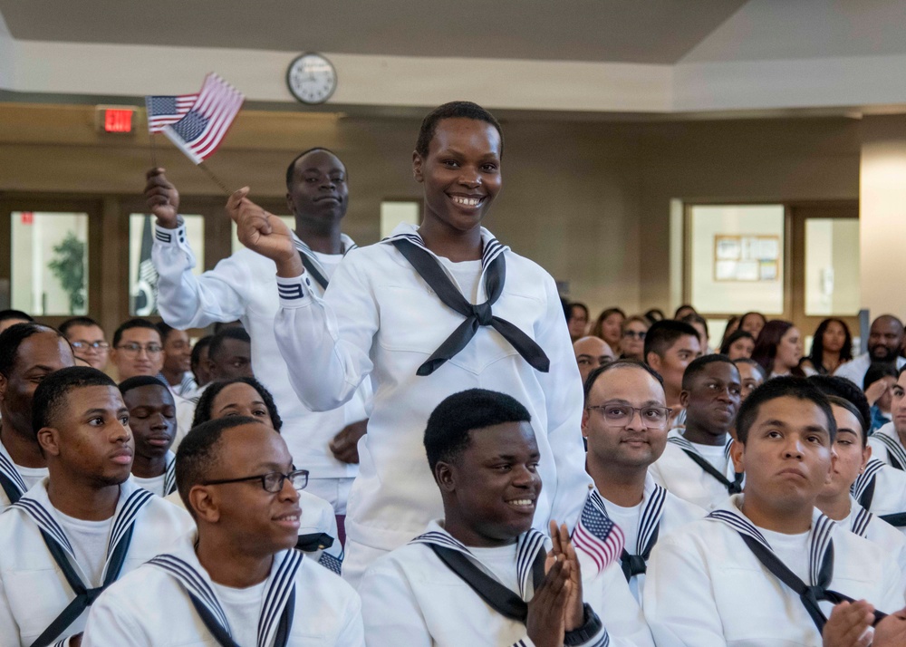 A group of U.S. Navy sailors in white dress uniforms are gathered in what appears to be an indoor ceremony hall. A central figure is standing and smiling broadly while waving a small American flag, with another sailor behind also waving a flag. The atmosphere appears celebratory, consistent with a naturalization or graduation ceremony.